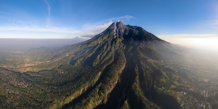 Aerial view of Mount Merapi with its lush forested slopes and deep ravines under a clear blue sky in Kemalang, Central Java, Indonesia.
