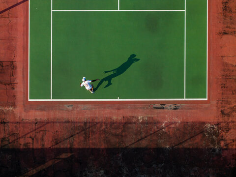 Aerial view of a tennis player celebrating a point on a green court with red borders, casting a long shadow in Kecamatan Prambanan, Jawa Tengah, Indonesia.