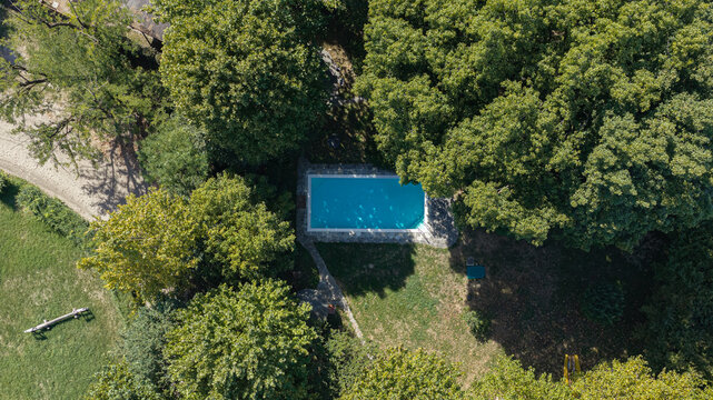 Aerial view of a rectangular swimming pool with bright blue water surrounded by lush green trees and a stone patio in Milan, Lombardy, Italy.
