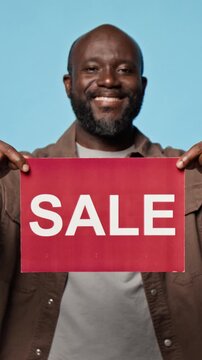 Portrait of smiling African American man holding red sale sign against blue background. Clip fits retail promo, shopping discount advertising, vertical