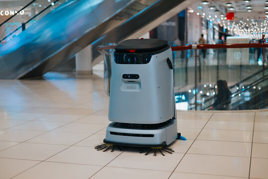A robot cleaning device stands on the tiled floor in a shopping mall. The escalators are visible in the background, indicating a busy indoor setting