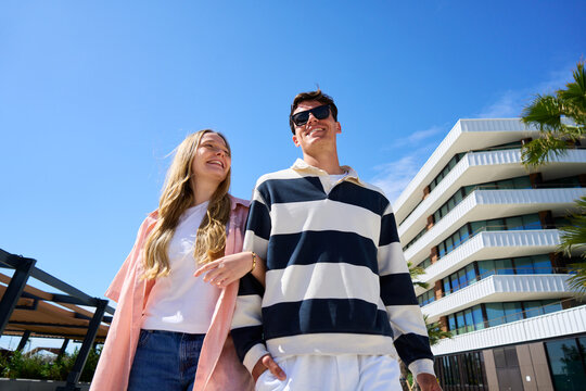Young couple enjoying a sunny day stroll, linking arms on a modern walkway.