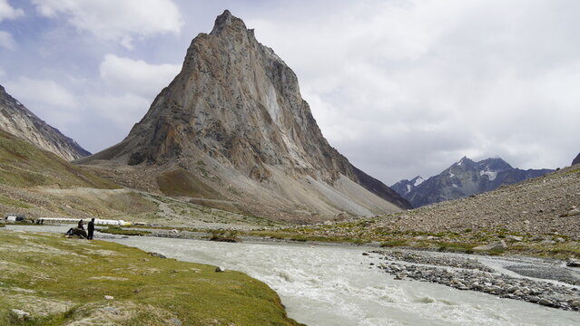 Rugged mountain peak and the mighty Gonbo Rangjon overlooking a river with people by the banks under cloudy skies in Zanskar Valley in Ladakh, India