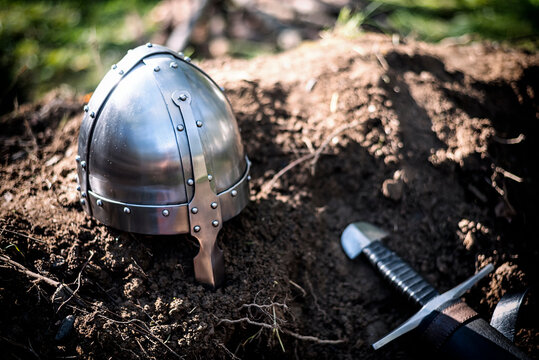 Primer plano de un casco spangenhelm vikingo o normando medieval de acero brillante y una espada descansando sobre tierra oscura y h&uacute;meda a plena luz del d&iacute;a.