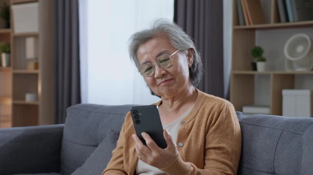 Smiling senior woman looking at smartphone while relaxing on sofa at home, enjoying digital technology, online connection and modern retirement lifestyle.