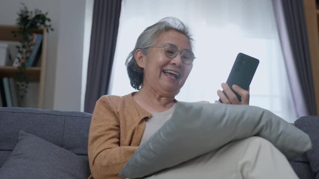 Joyful senior woman laughing while looking at smartphone at home, enjoying digital entertainment, online connection and modern technology during retirement