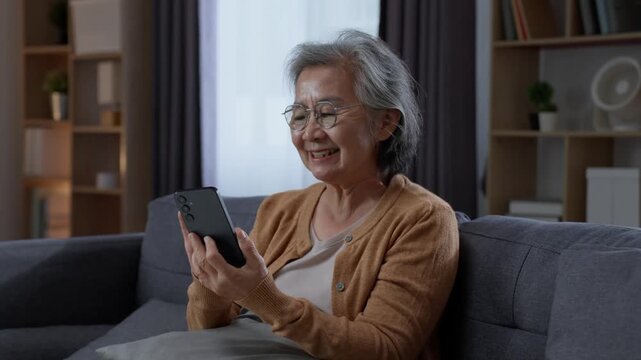Cheerful senior woman laughing while looking at smartphone at home, enjoying entertaining digital content and modern mobile technology during her retirement leisure time