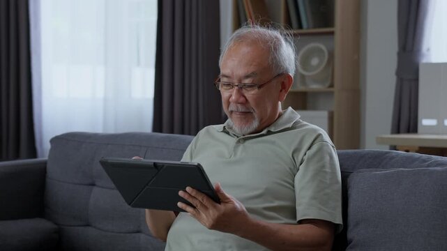 Senior man looking at digital tablet while sitting comfortably on sofa at home, browsing online content and using modern technology during his everyday retirement life