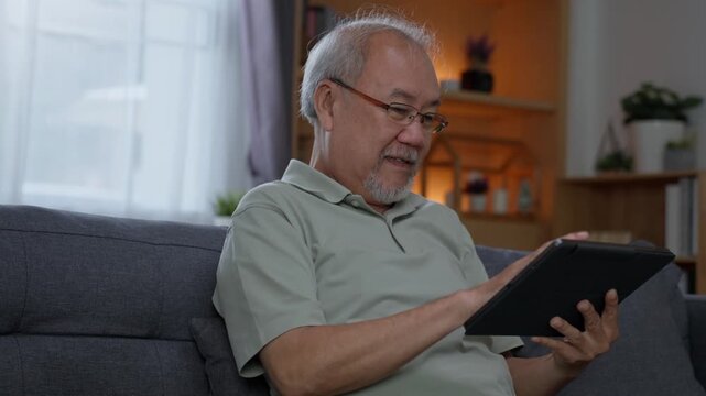 Senior man looking at digital tablet while relaxing on sofa at home, reading and browsing online content with modern technology during his everyday retirement lifestyle.