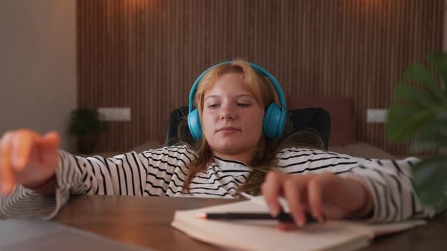 Teenage student distracted from homework while listening to music through headphones at home, studying, representing procrastination, distraction and lack of productivity