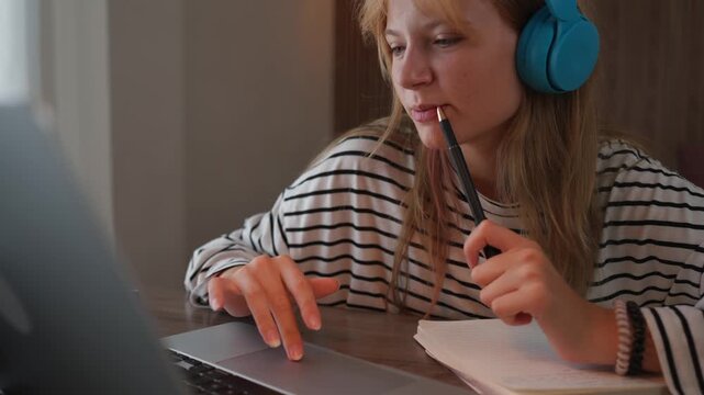 Teenage girl wearing headphones participating in an online lesson, looking at her laptop screen and listening to the teacher while thoughtfully chewing on a pen during her e-learning session