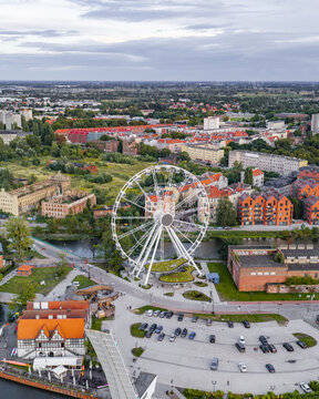 Aerial view of the AmberSky ferris wheel situated near the river, surrounded by historic and modern buildings, green spaces, and a parking area in Gdansk, Pomeranian Voivodeship, Poland.