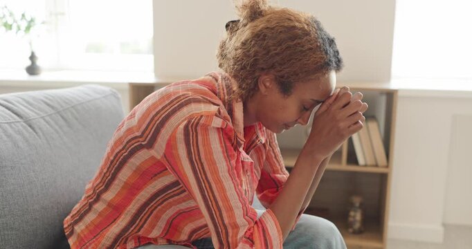 Stressed woman sits on sofa, feeling anxious. Adult bows her head with clasped hands in a sunlit living room, expressing worry, sadness and exhaustion. Mental health struggle at home.