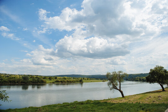 Scenic view of the calm Dniester River bank in Soroca, Moldova, featuring green trees along the shore and a bright blue sky with large fluffy clouds.