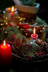 Fototapeta premium Dark and cozy Christmas arrangement with red candles, evergreen branches, red berries and pinecones. Warm candlelight and soft bokeh create a moody, intimate festive atmosphere.