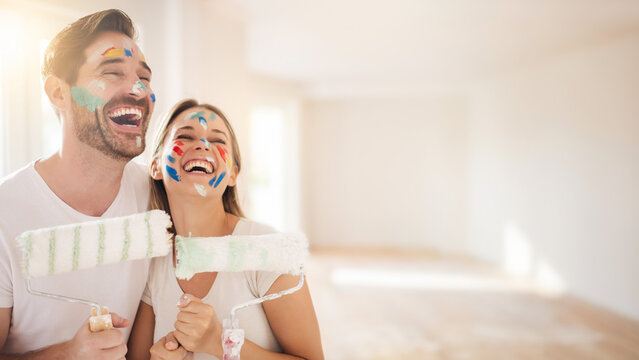 Playful young couple with paint on faces holding rollers with blurred sunny apartment background banner.