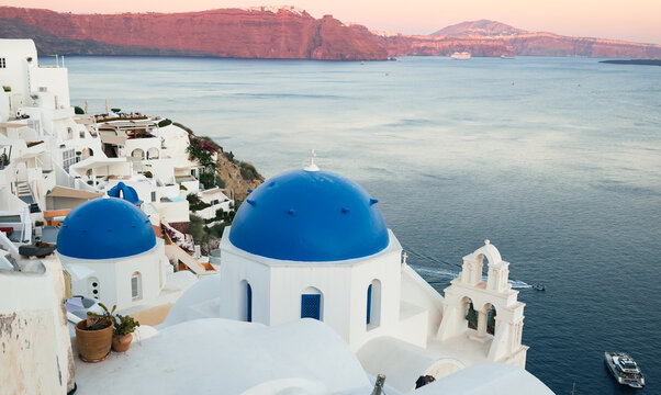 View of Santorini, Greece with blue dome churches and cliff views of houses and businesses overlooking the caldera. Famous tourism destination.