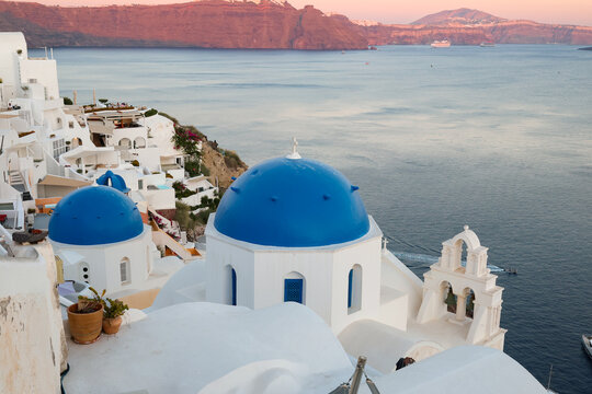 View of Santorini, Greece with blue dome churches and cliff views of houses and businesses overlooking the caldera. Famous tourism destination.