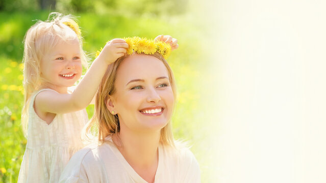 Happy little girl placing flower crown on mother with blurred spring meadow background banner.