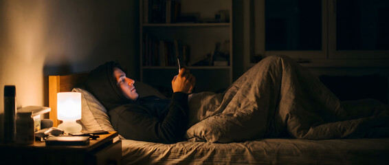 Fototapeta premium An adult man lies in a dimly lit bedroom at night, using a mobile phone while resting in bed.