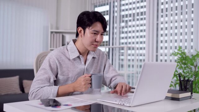 Young Asian man holding coffee mug typing on laptop working hybrid at home office desk