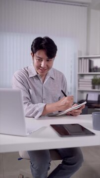 Smiling Asian man writing notes while video calling on laptop at home office desk