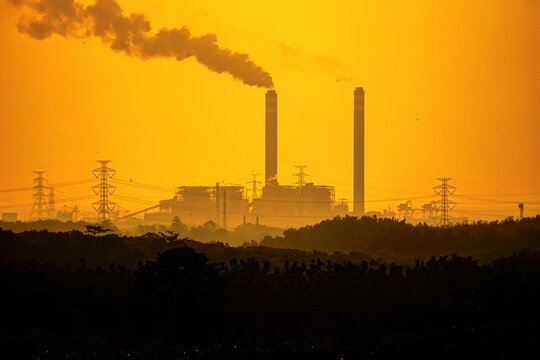 Telephoto view of industrial plant chimneys blowing thick smoke against a bright yellow sunset sky.