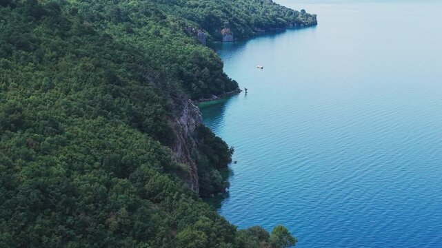 Drone view of a scenic road winding along rocky, forested cliffs and the turquoise waters of Lake Ohrid near Ohrid, North Macedonia. Natural shoreline, secluded beach and clear water on a sunny summer