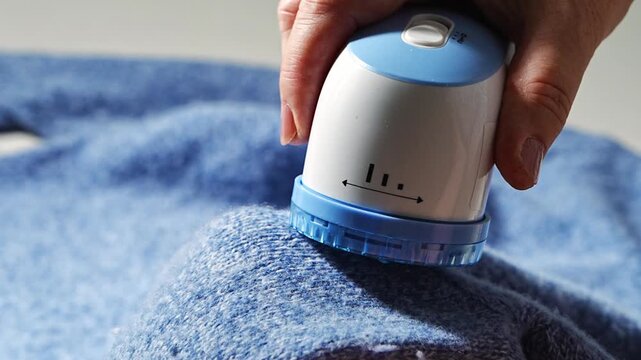 Woman removing fabric pills, lint from knitted wool sweater with an electric shaver. Close up hands on process, garment care, reuse, durability.
