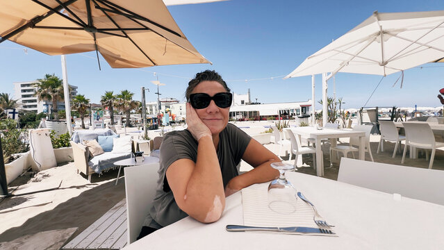 A woman wearing sunglasses sitting at an outdoor restaurant table near a beach.