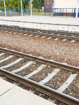 Parallel railway tracks with steel rails, concrete sleepers, gravel ballast, and platform edge beside fenced urban station infrastructure