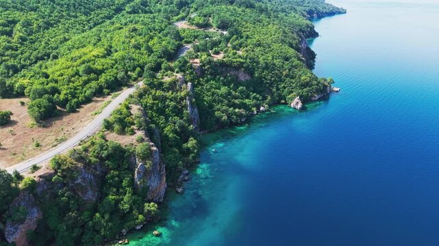 Drone view of a scenic road winding along rocky, forested cliffs and the turquoise waters of Lake Ohrid near Ohrid, North Macedonia. Natural shoreline, secluded beach and clear water on a sunny summer