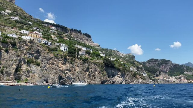 Picturesque view from sailing yacht on amazing rock hill with old scattered houses along the Amalfi coast in sunny day, turquoise water with wake foam, rocking boats and sailing seagulls
