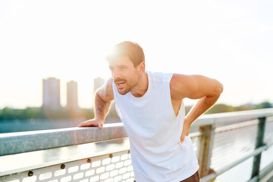 Male athlete suffering from lower back pain while leaning on a railing after intense exercise on a bridge in the city