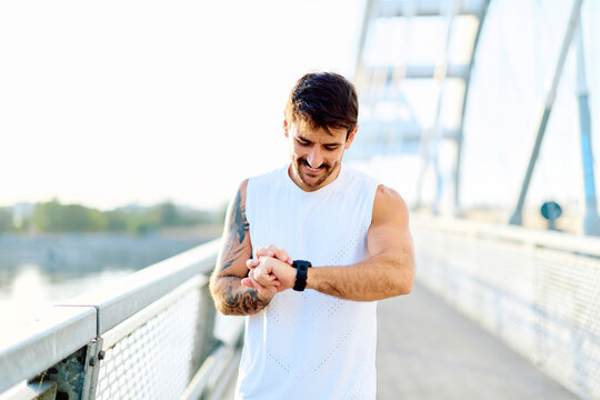 Smiling runner athlete checking performance data on a wearable device while training on a city bridge
