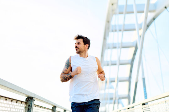 Man with tattoos running on a modern urban bridge, focusing on fitness and a healthy lifestyle. He is exercising outdoors