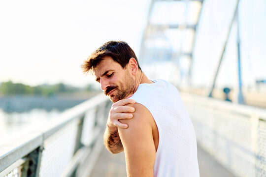 Male athlete suffering from shoulder injury while running outdoors on a bridge, experiencing discomfort and muscle pain