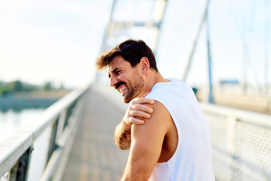 Male runner suffering from shoulder pain and injury, grimacing during an outdoor workout on a bridge, depicting muscle strain