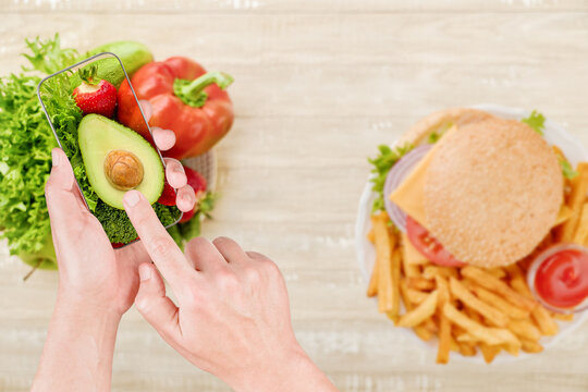 Hands use a phone over vegetables while a burger sits on a wooden table