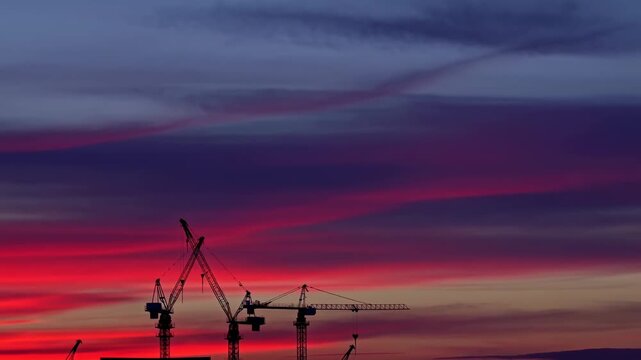 Construction cranes operate over an urban building site beneath layered sunset clouds. Construction industry advertising, real estate development marketing, and infrastructure presentations.