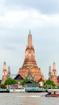Wat Arun temple standing beside the Chao Phraya River in Bangkok, Thailand, with a traditional long-tail boat crossing the water under a bright cloudy sky, creating a scenic travel view.