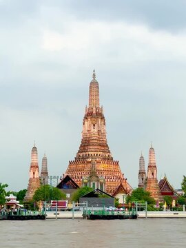 Wat Arun temple standing beside the Chao Phraya River in Bangkok, Thailand, with a traditional long-tail boat crossing the water under a bright cloudy sky, creating a scenic travel view.