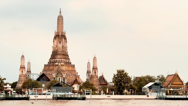 Wat Arun temple standing beside the Chao Phraya River in Bangkok, Thailand, with a traditional long-tail boat crossing the water under a bright cloudy sky, creating a scenic travel view.