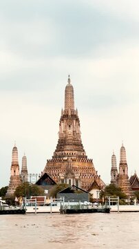 Wat Arun temple standing beside the Chao Phraya River in Bangkok, Thailand, with a traditional long-tail boat crossing the water under a bright cloudy sky, creating a scenic travel view.