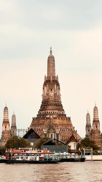 Wat Arun temple standing beside the Chao Phraya River in Bangkok, Thailand, with a traditional long-tail boat crossing the water under a bright cloudy sky, creating a scenic travel view.