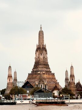 Wat Arun temple standing beside the Chao Phraya River in Bangkok, Thailand, with a traditional long-tail boat crossing the water under a bright cloudy sky, creating a scenic travel view.