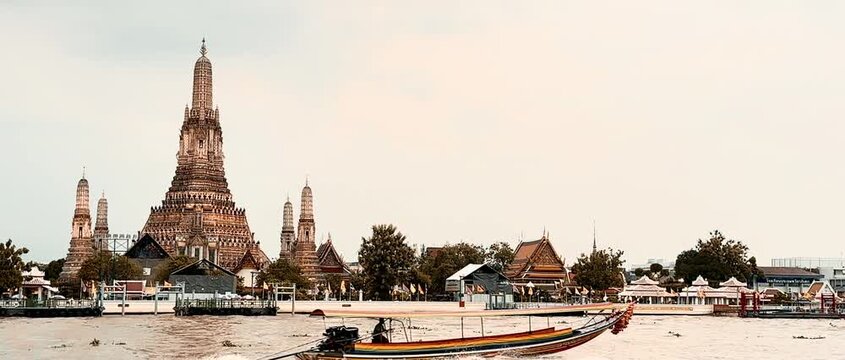 Wat Arun temple standing beside the Chao Phraya River in Bangkok, Thailand, with a traditional long-tail boat crossing the water under a bright cloudy sky, creating a scenic travel view.