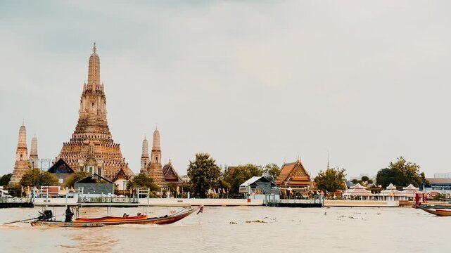 Wat Arun temple standing beside the Chao Phraya River in Bangkok, Thailand, with a traditional long-tail boat crossing the water under a bright cloudy sky, creating a scenic travel view.