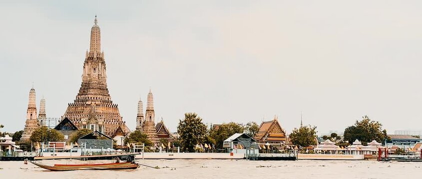 Wat Arun temple standing beside the Chao Phraya River in Bangkok, Thailand, with a traditional long-tail boat crossing the water under a bright cloudy sky, creating a scenic travel view.