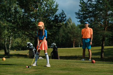 Obraz premium Young girl practices golf swing on a sunny day while an adult watches on a lush green course surrounded by trees in the background